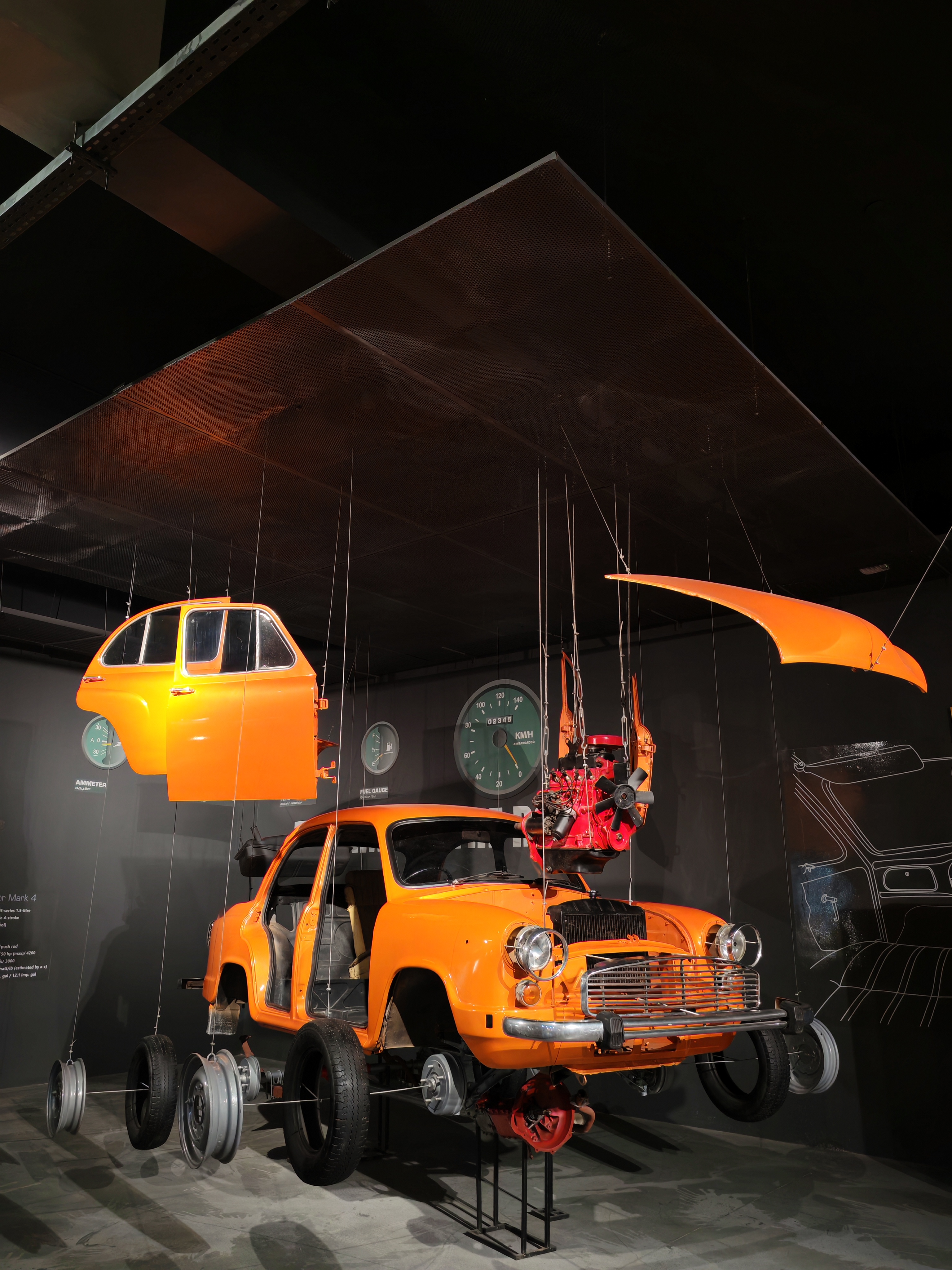 An orange Ambassador car installation suspended in a dark gallery